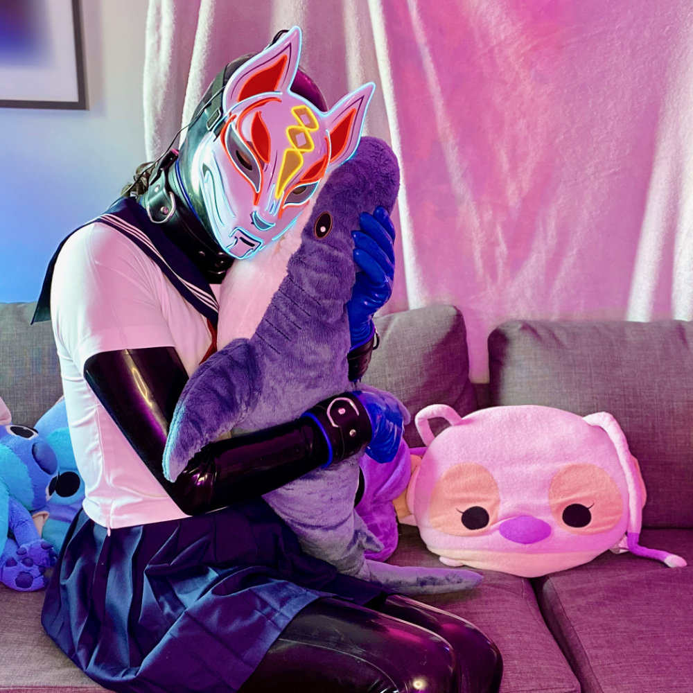 Black latex catsuit with blue gloves, toe socks, and restraints. Wearing a white and blue Japanese seifuku with a light-up Japanese fox mask. Hugging IKEA plushie shark