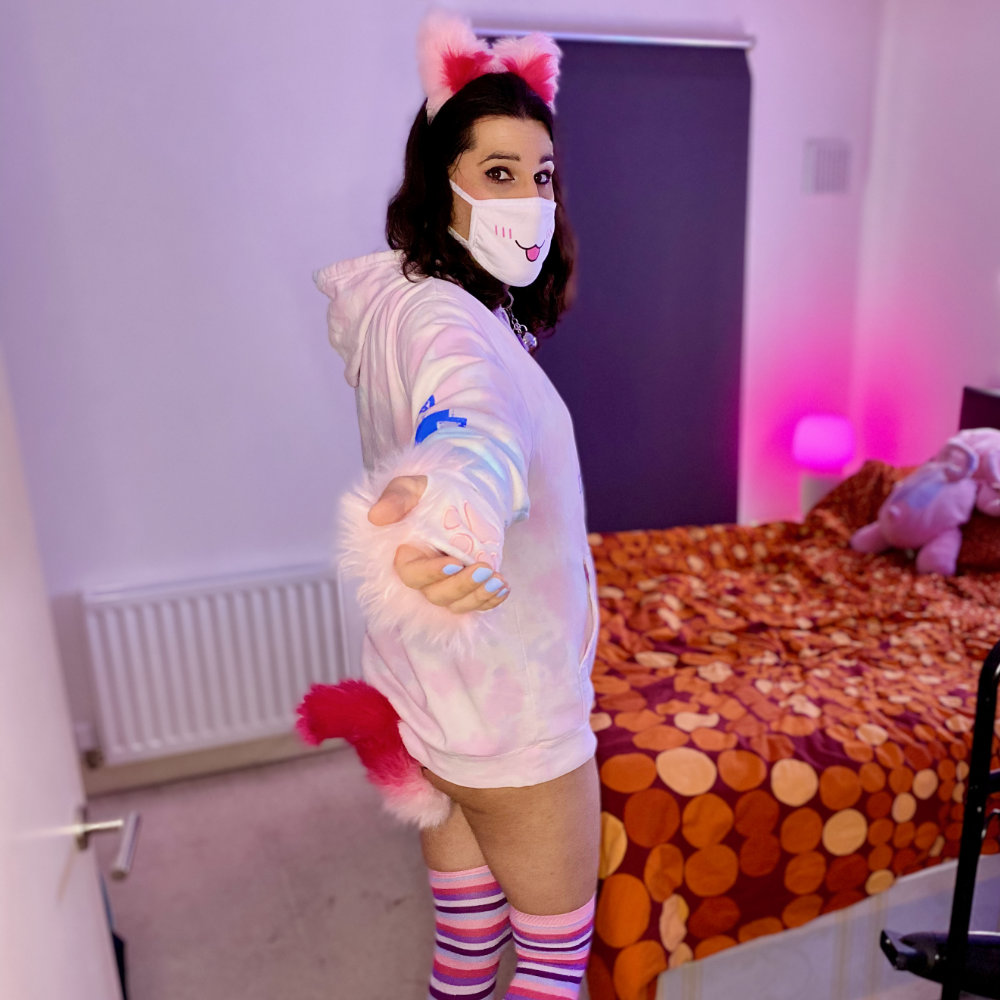 Me beckoning you to come into the bedroom, wearing pink cat ears, red and white tail plug, pink and blue hoodie, pastel blue nails, stripy pink knee-high toe socks