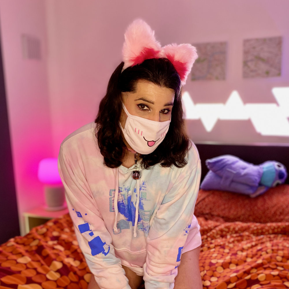 Me in pastel pink and blue hoodie, wearing pink cat ears, collar with bell, paws, and toe socks. Kneeling on bed.