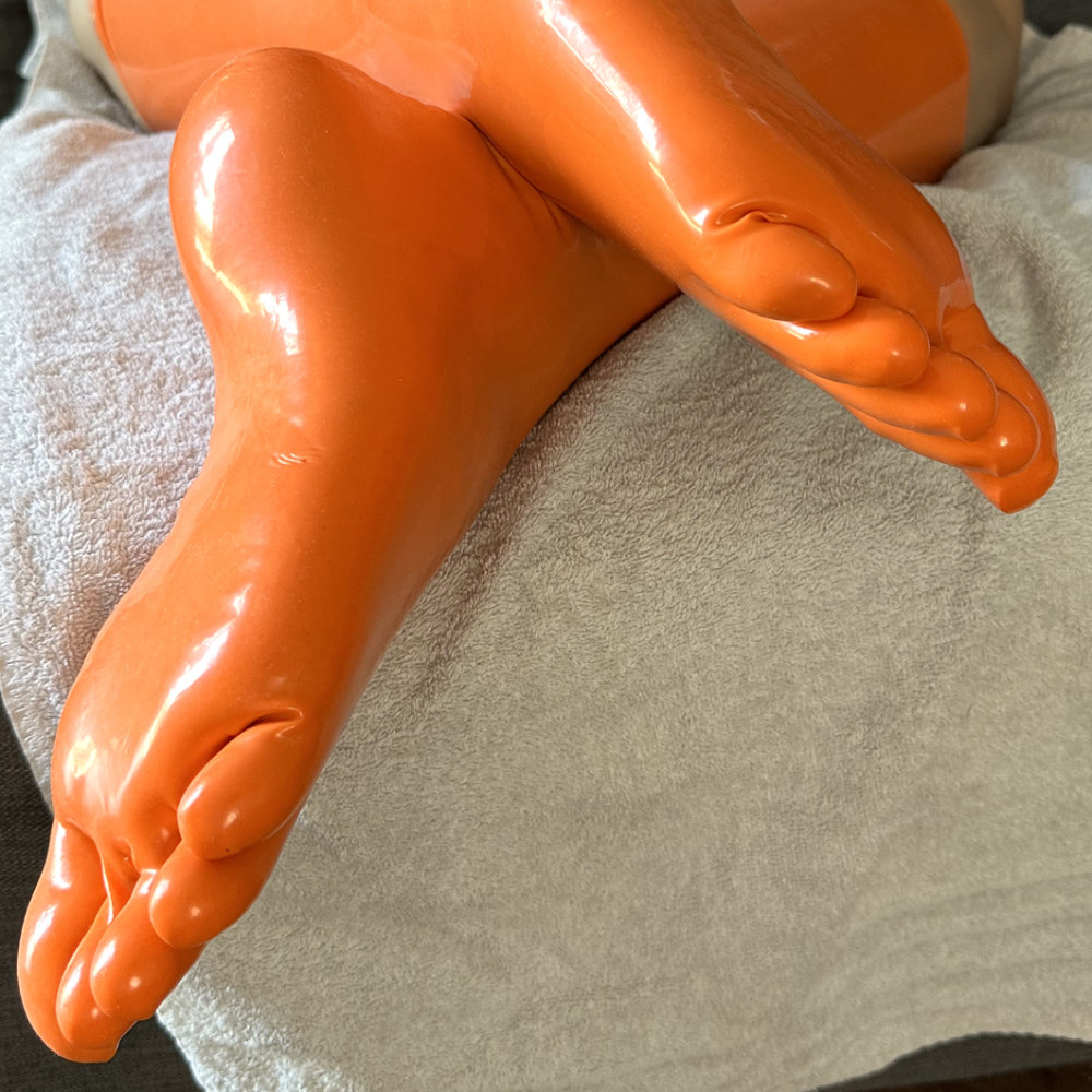 My feet in shiny knee-high orange latex toe socks. I am kneeling on the sofa, feet crossed at the ankle, with soles visible to camera