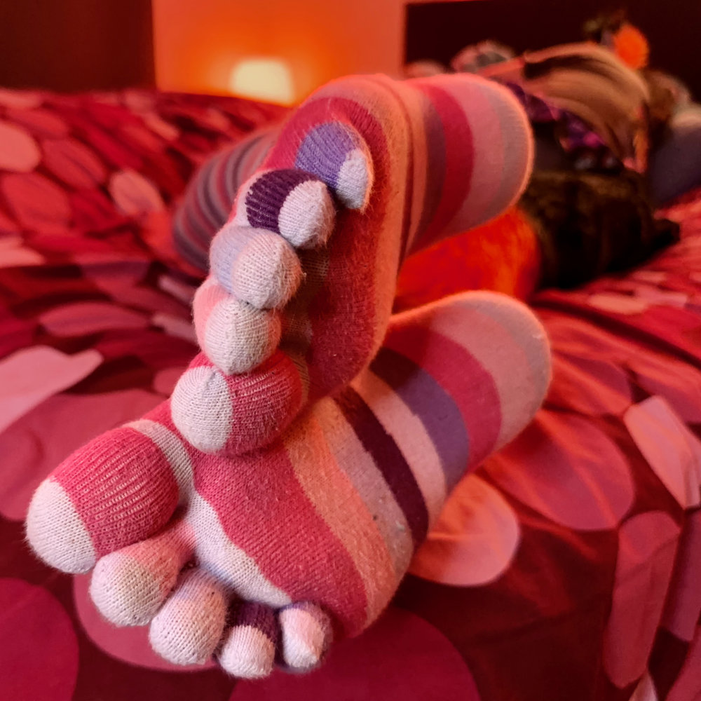 Rina's feet in knee-high, stripy purple and pink toe socks, with the toes and soles visible to camera. Rina is lying on her bed facing away from the camera in the background