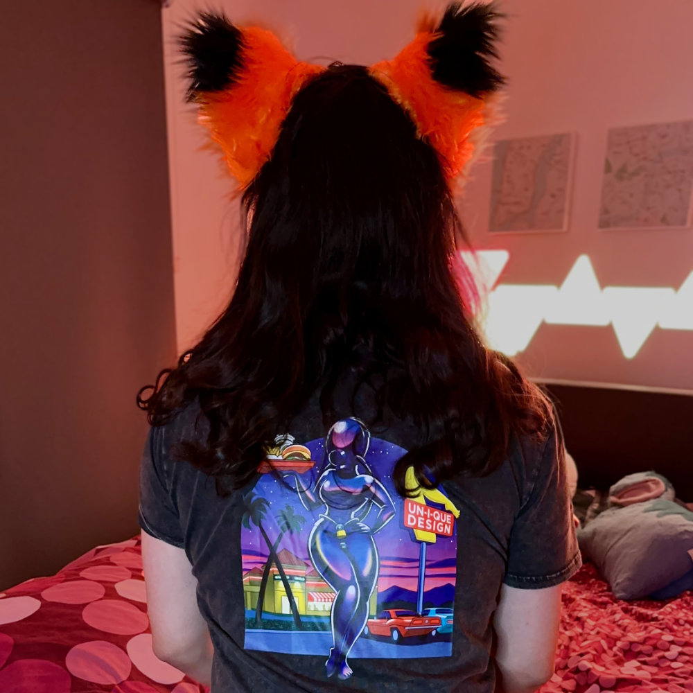Rina facing away from camera, showing the design on the back of her t-shirt. It’s by UniqDsn, showing a rubber doll holding a fast food tray in a 50s, In-n-Out Burger inspired-design. Rina is also wearing a purple plaid miniskirt, a black fox tail, black and orange fox ears, and stripy toe socks