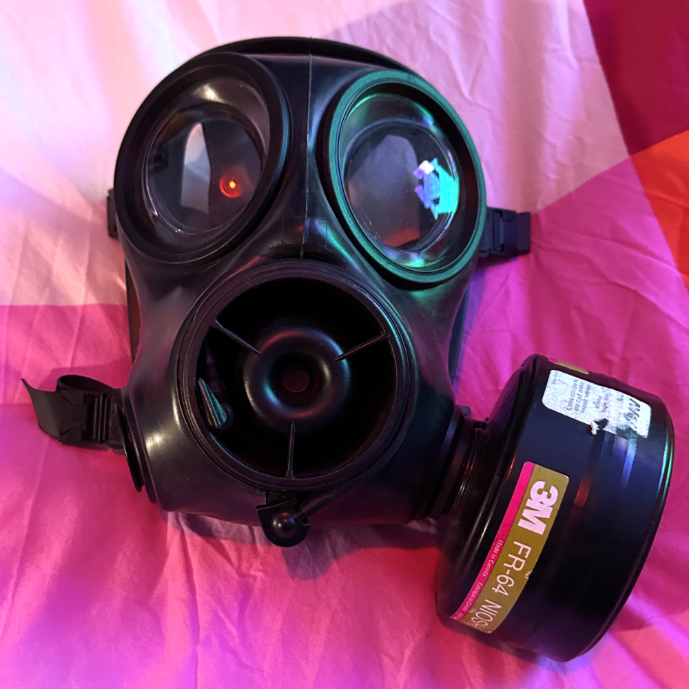 A black S10 gas mask, with clear lenses and a filter attached, resting on a bedsheet. The ambient colour is mostly pink, with a greenish-blue cyber fox-shaped light reflecting in the lens