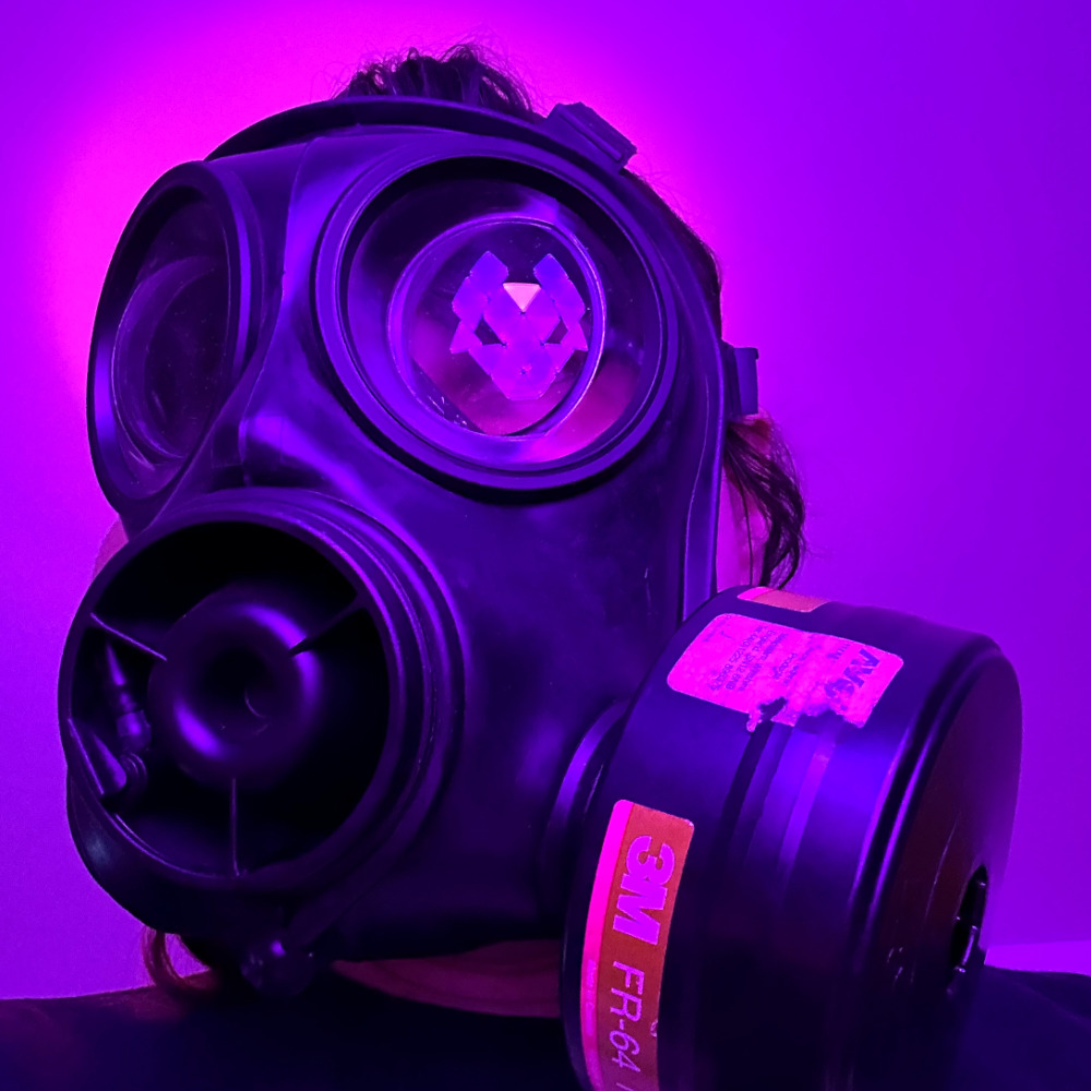 Rina wearing a black S10 gas mask, looking past the left hand side of the camera. In the mask’s right lens is a reflection of the cyber fox light, glowing purple. Behind my head is a halo-like light effect glowing pink