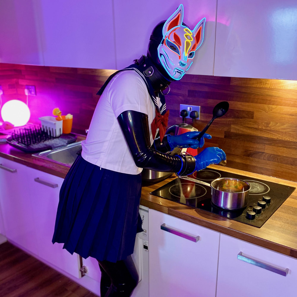 Black latex catsuit, blue latex gloves and toe socks, fox mask, Japanese seifuku. Adding curry roux to a pot.