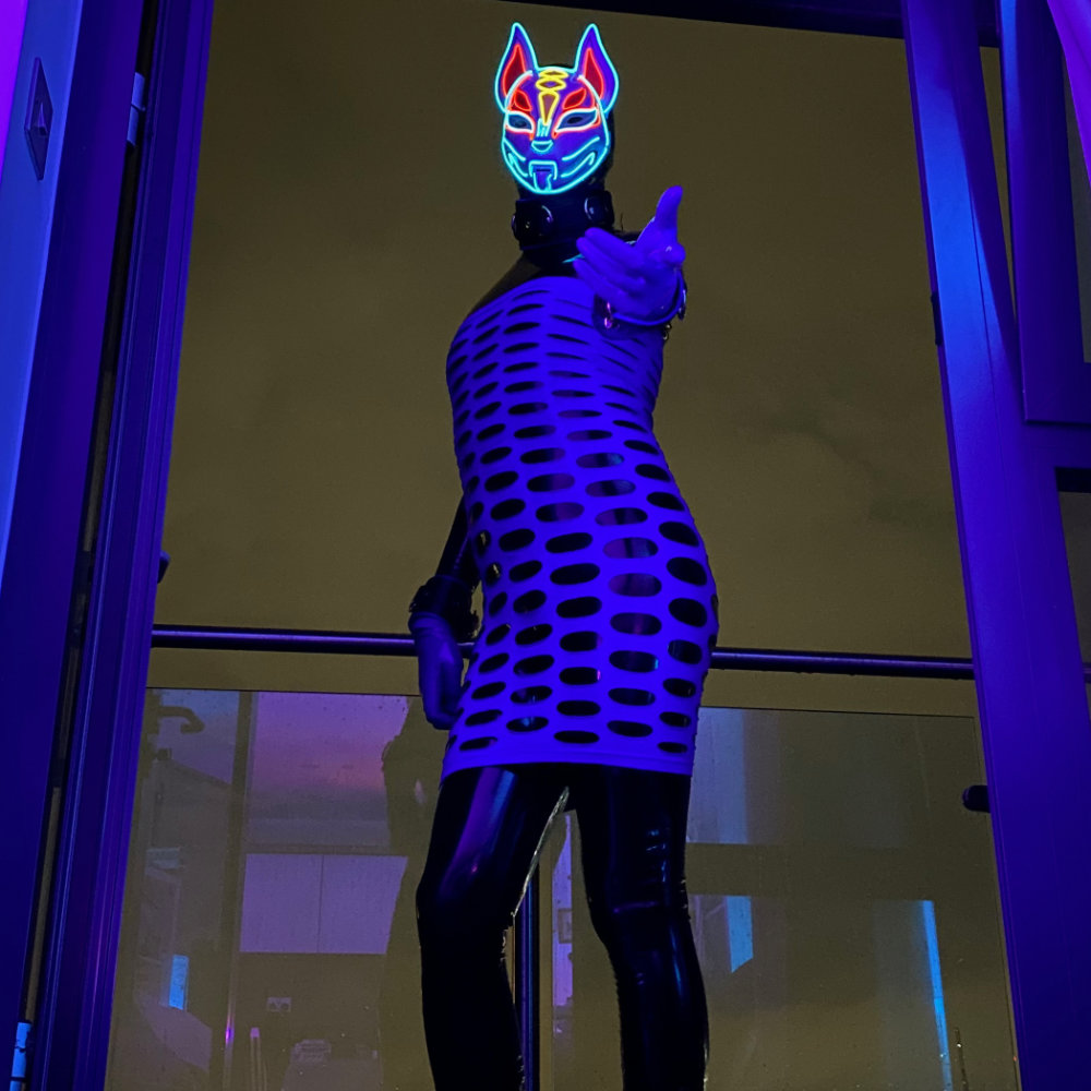 Black latex catsuit with purple hole dress, and purple gloves & toe socks. Wearing a light-up  Japanese fox mask. Standing on balcony at night, beckoning to camera