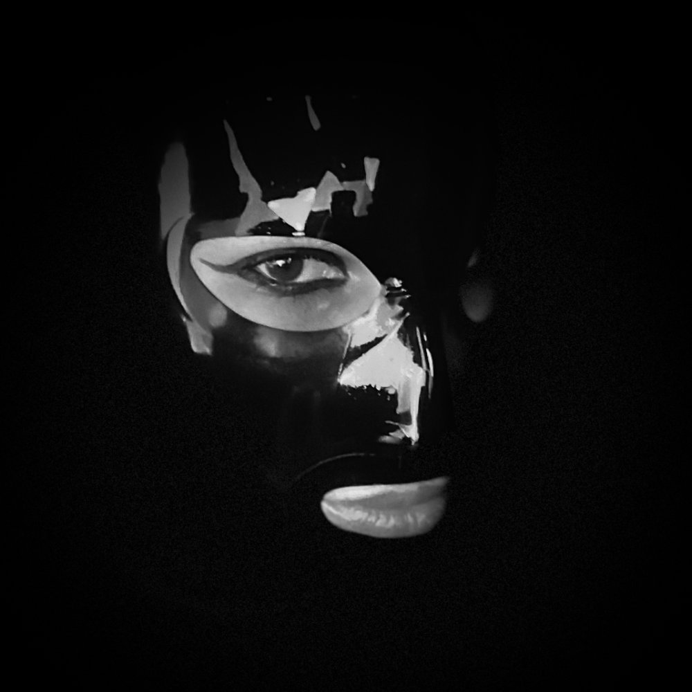 Black & white photo of my latex-hooded face, surrounded by darkness, features lit up by the light reflecting in the shiny latex