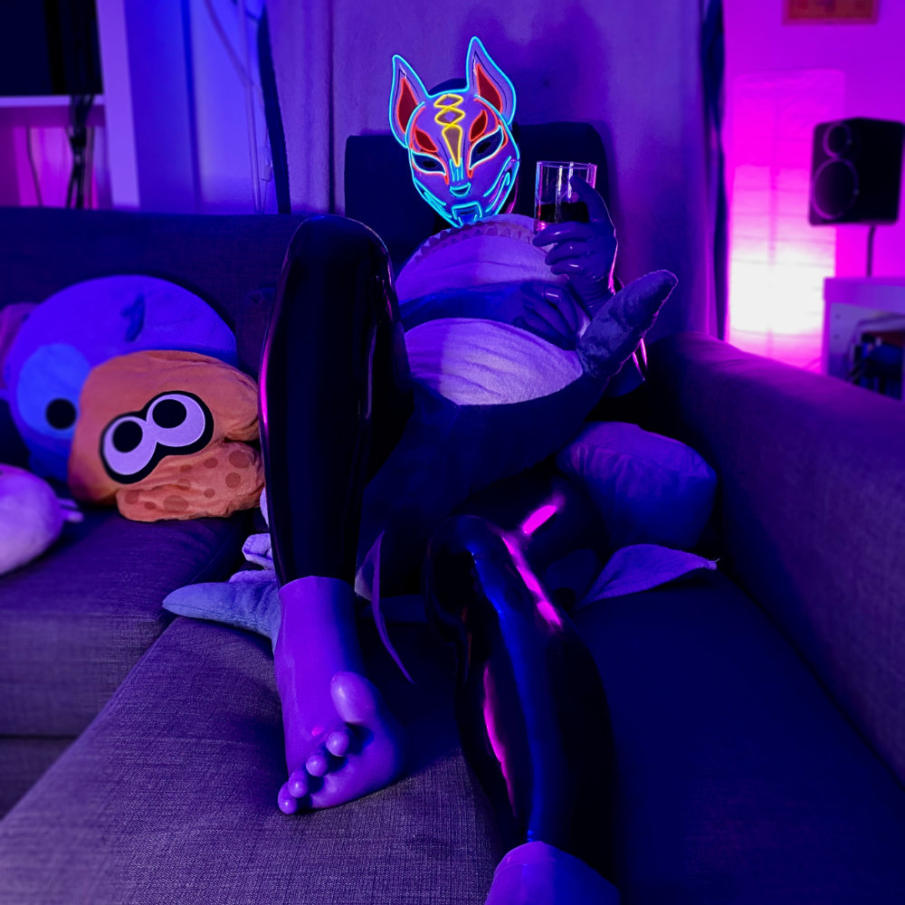 Lying on sofa dressed in black latex, with purple toe socks and gloves, and a light-up fox mask. Holding a drink in my left hand and hugging a shark plushie with my right