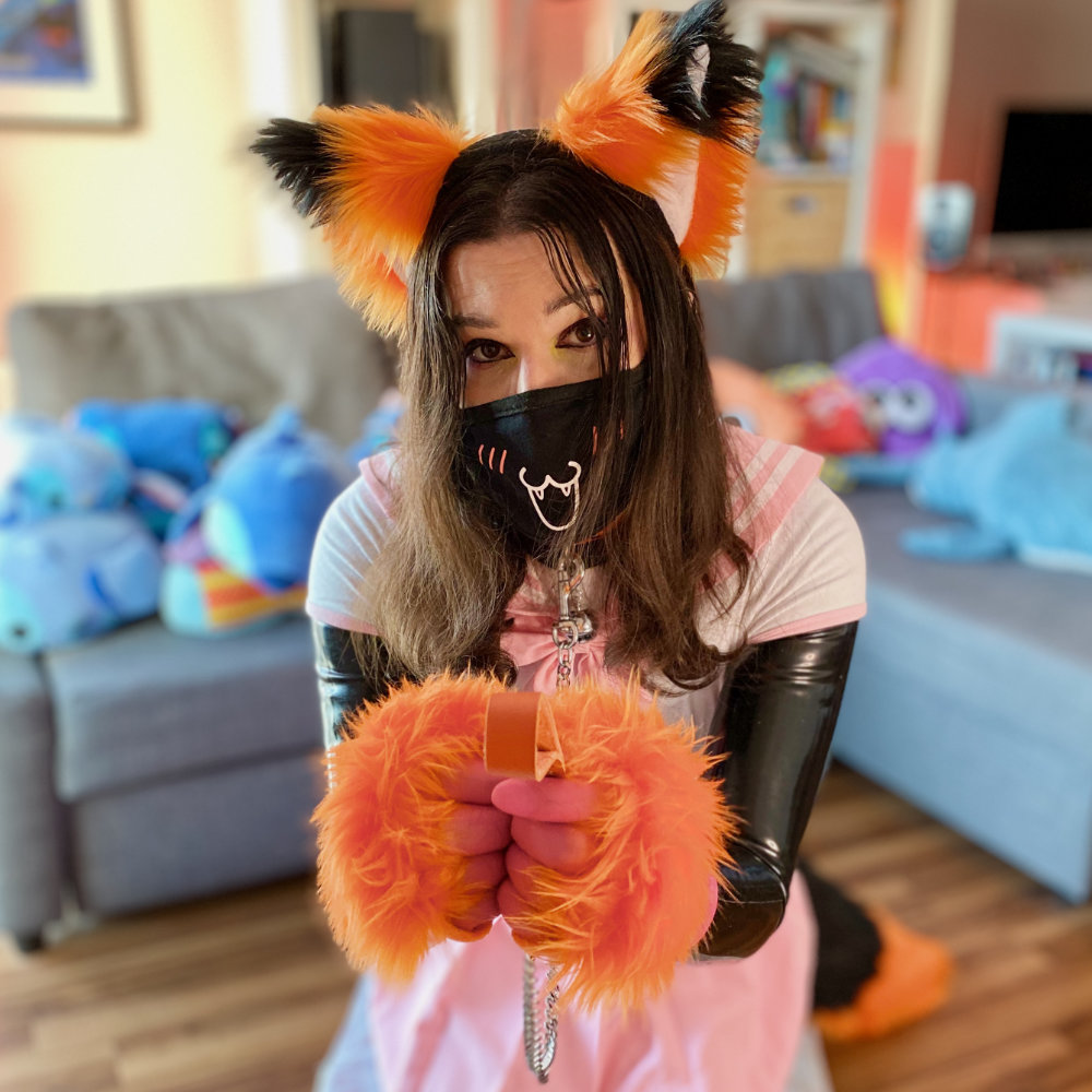 Rina kneeling on floor, offering her collar and leash to the camera. She is wearing a pink and white Japanese schoolgirl uniform over a black latex catsuit, with orange and black fox ears, paws, and tail