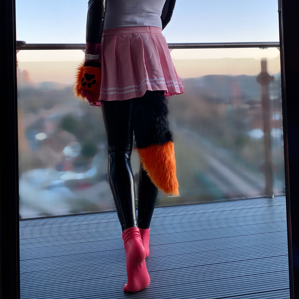 Rina standing on a high balcony, looking away from camera, and wearing a black latex catsuit with pink latex toe socks and gloves under a pink and white Japanese schoogirl uniform, and orange & black fox ears, paws, and tail